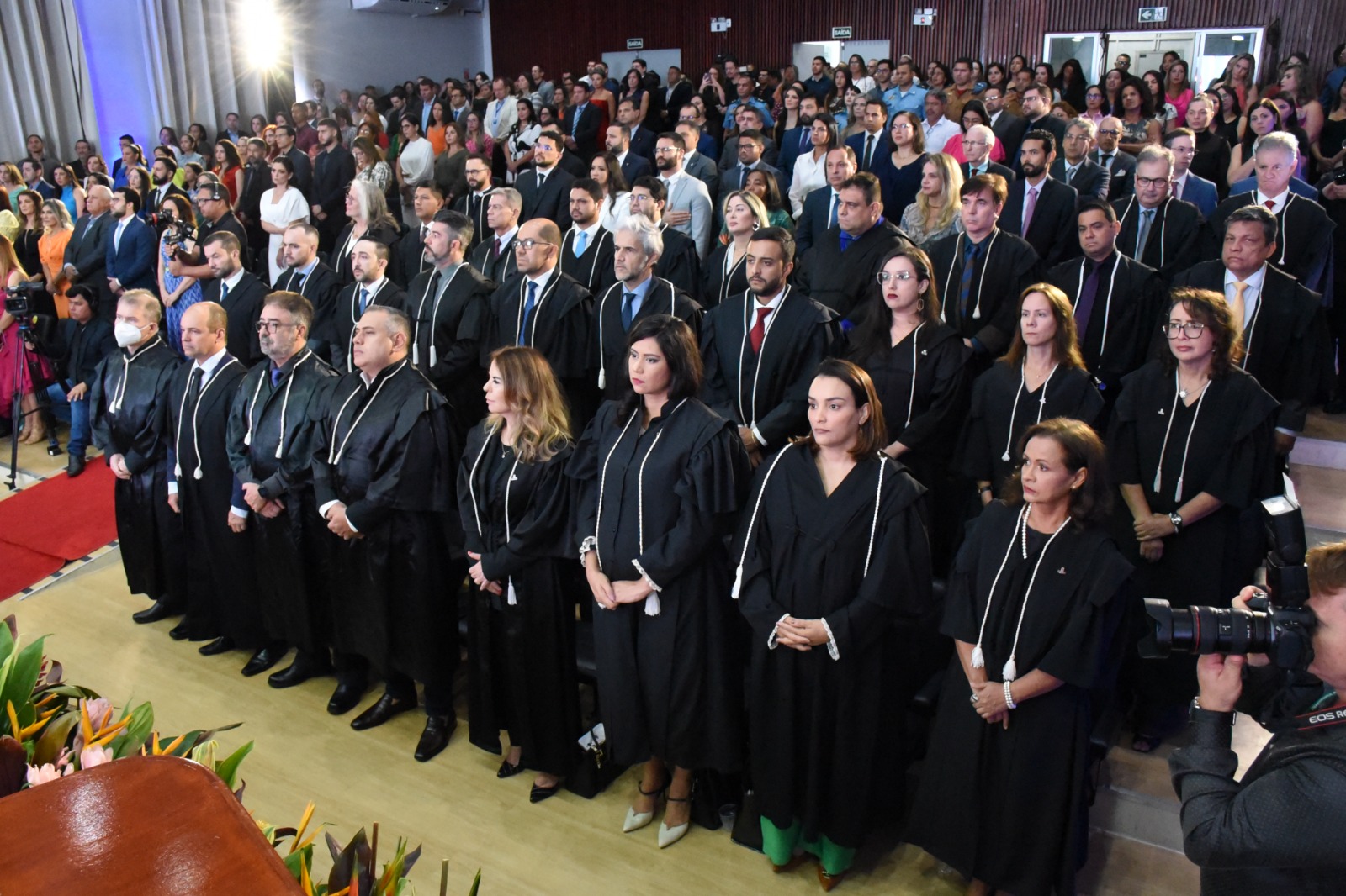 Foto colorida mostra pessoas em pé em um auditório.