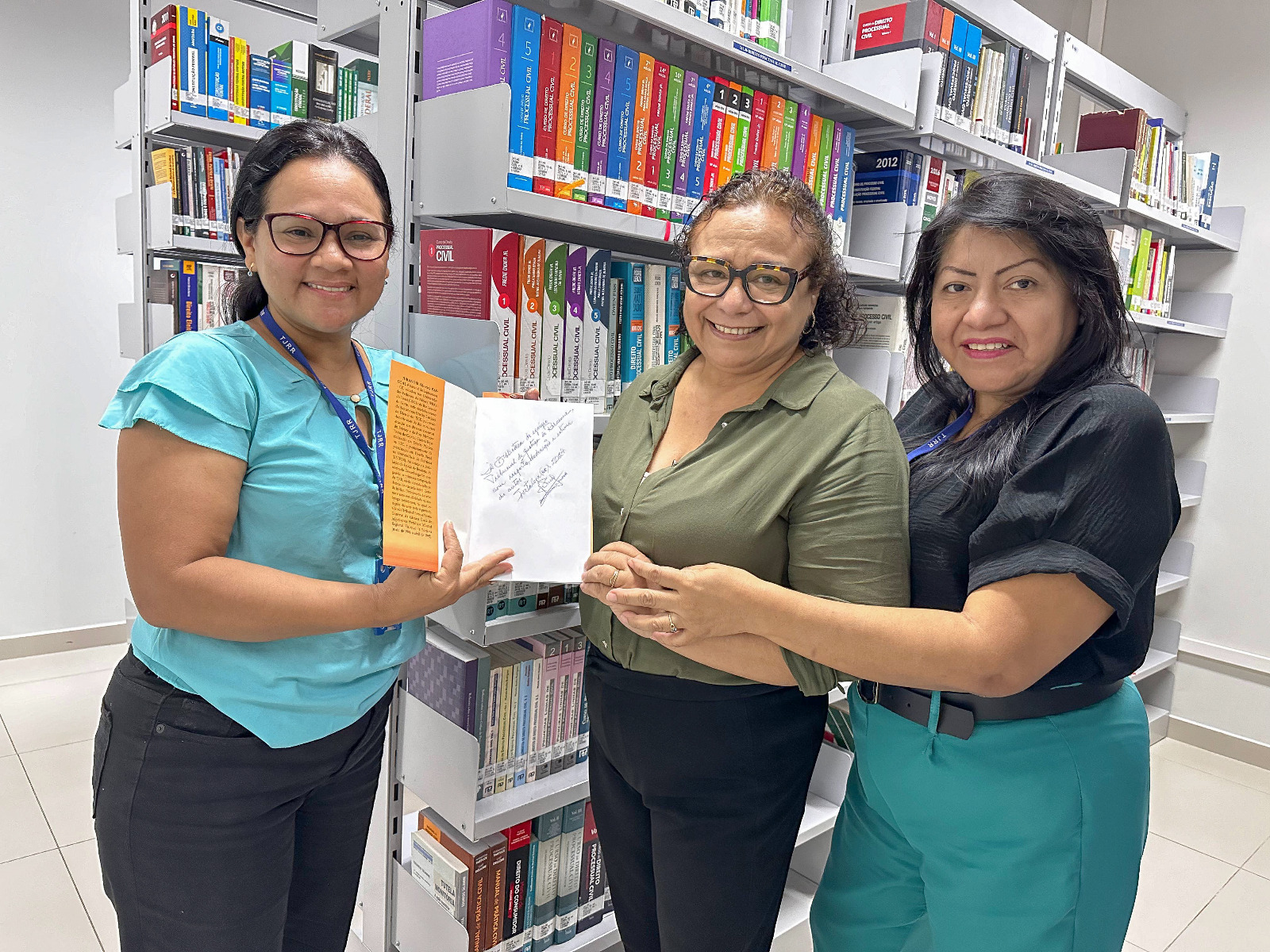 foto colorida de três servidores da biblioteca pousando para foto com o livro do desembargador Jurandir pascoal em mãos. O ambiente é interno, em uma biblioteca