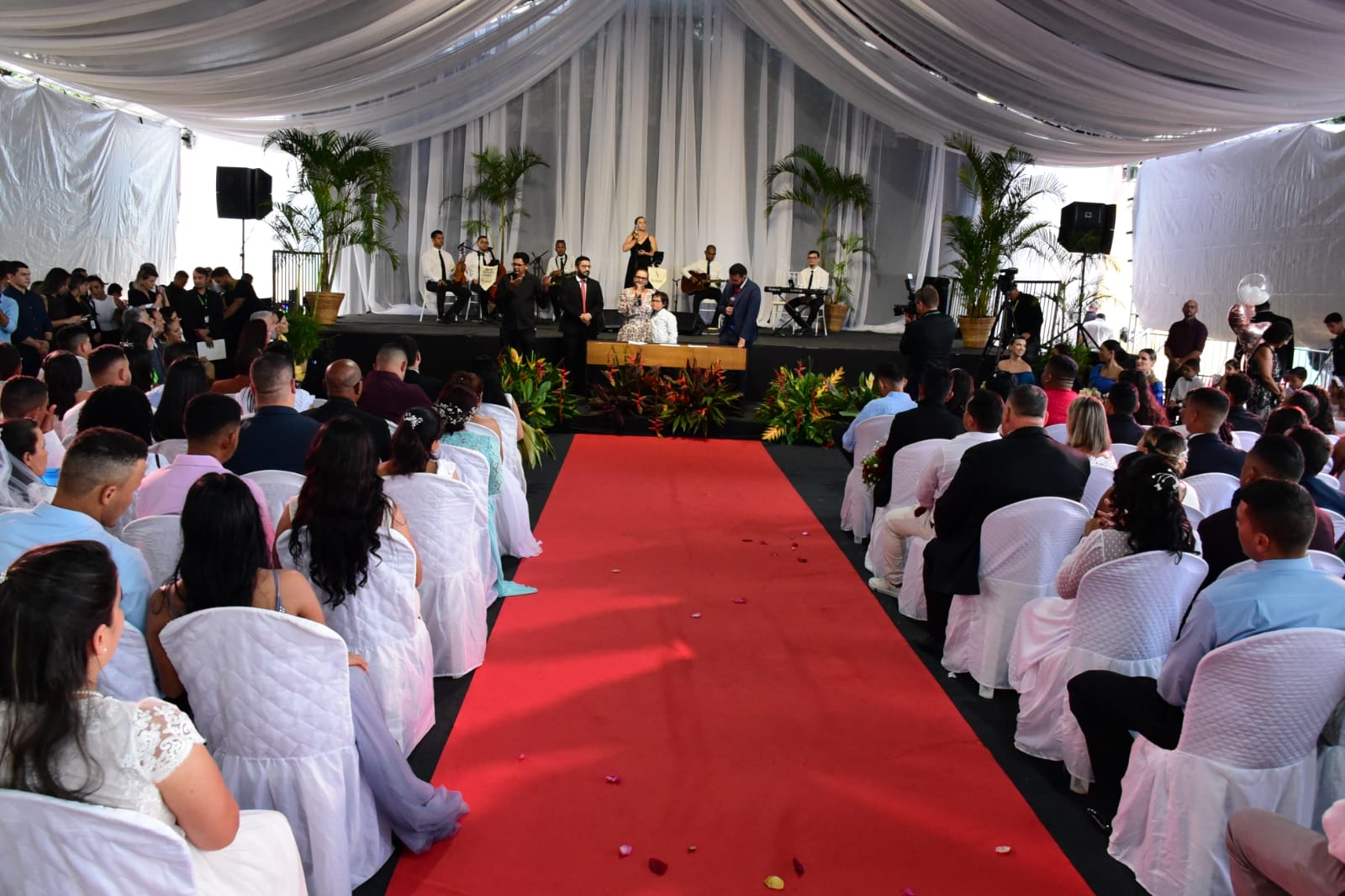 Foto colorida mostra aproximadamente 100 pessoas durante a cerimonia do casamento coletivo