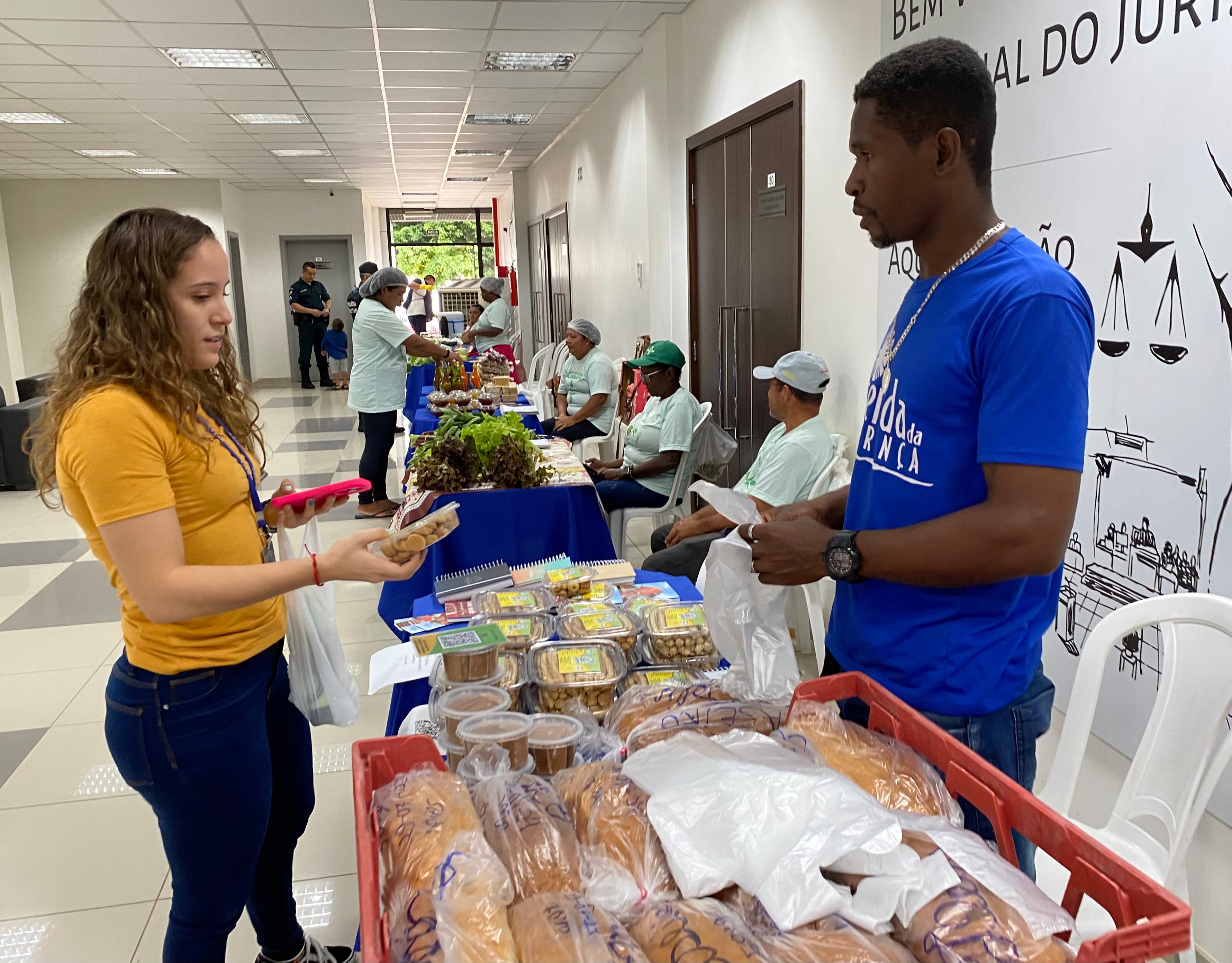 Imagem colorida mostra Feira de ôrganicos do TJRR, ocorrendo no Fórum Criminal Ministro Evandro Lins e Silva, com a participação das Camponesas do Lavrado. Na imgem é possivel ver servidora do TJRR fazendo pagando pela compra na feira.