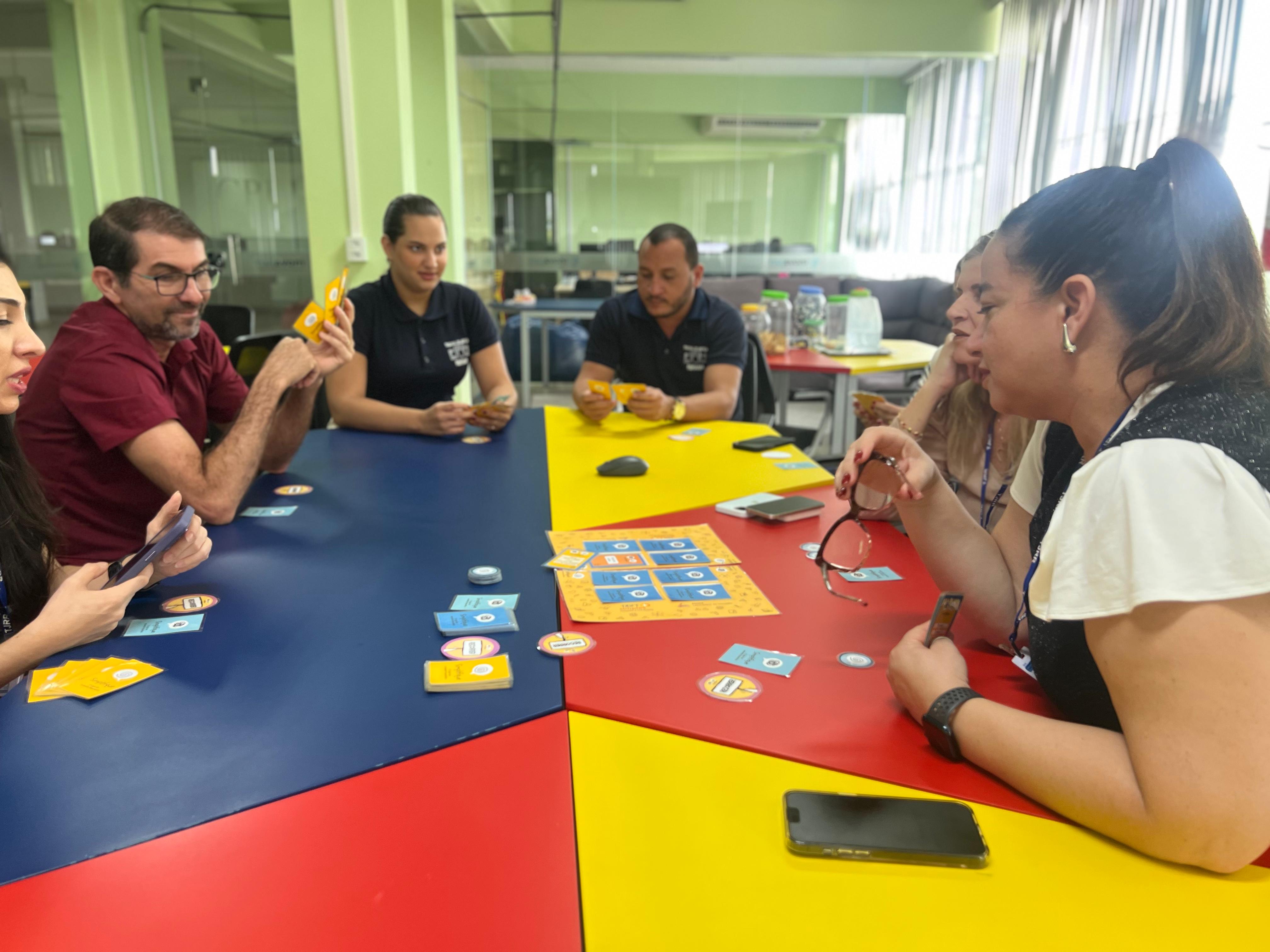 Imagem colorida mostra seis pessoas, entre homens e mulheres em sentados em volta de uma mesa colorida jogando jogo de narrativa simplificada. O jogo é em formato de baralho, contendado cartas e fichas.