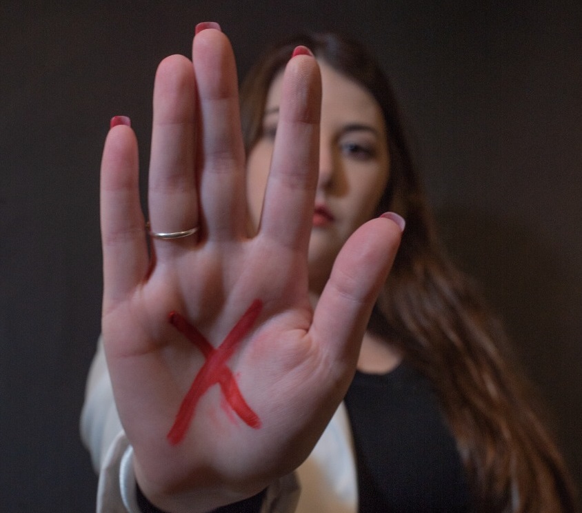 Imagem colorida de mulher com mão estendida para frente do corpo com a palma da mão aberta, contendo um “x” feito de caneta vermelha. 