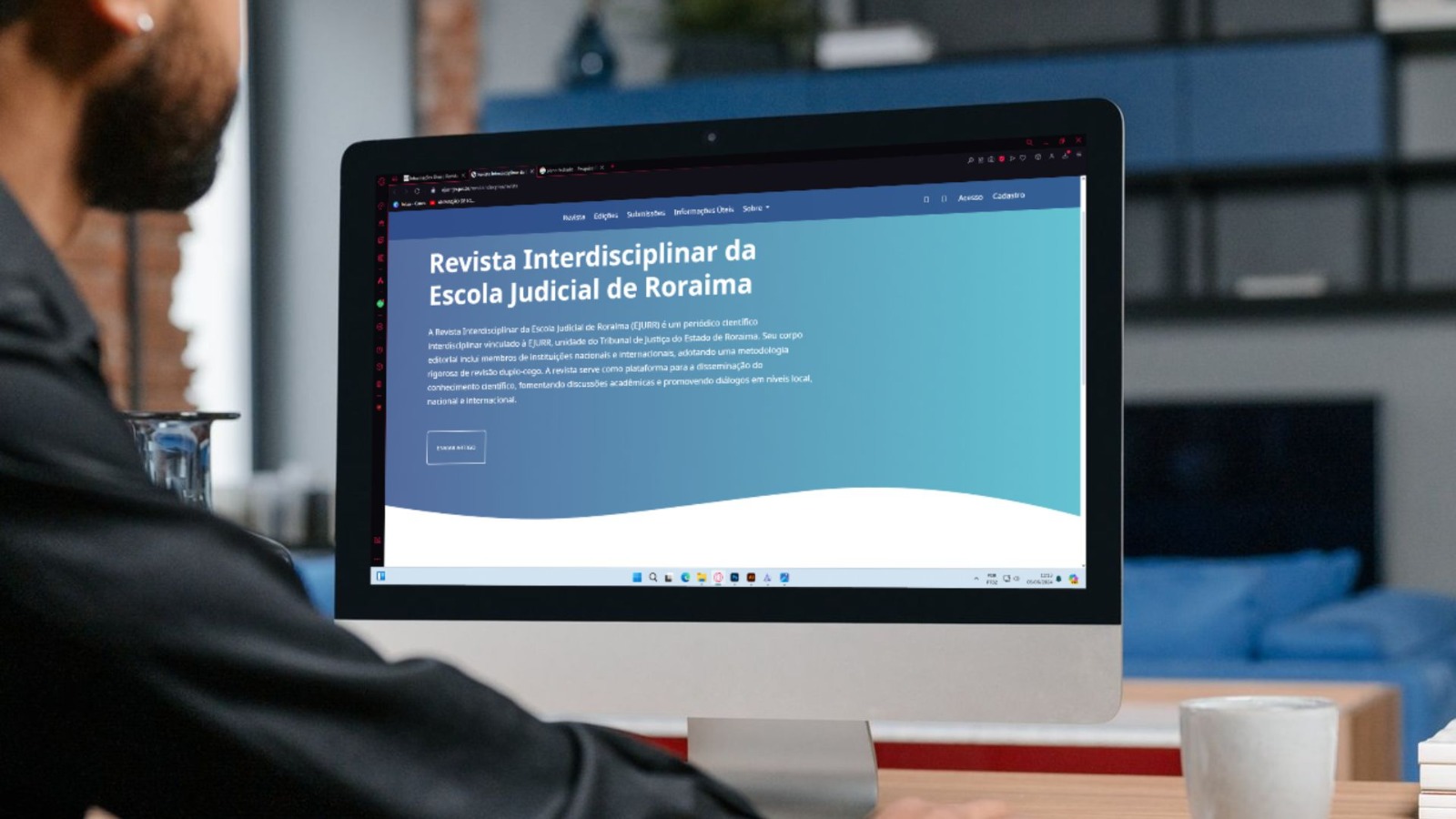 Imagem colorida mostrando um homem sentado em frente a um computador. Na tela do computador o texto: Revista Interdisciplinar da Escola Judicial de Roraima