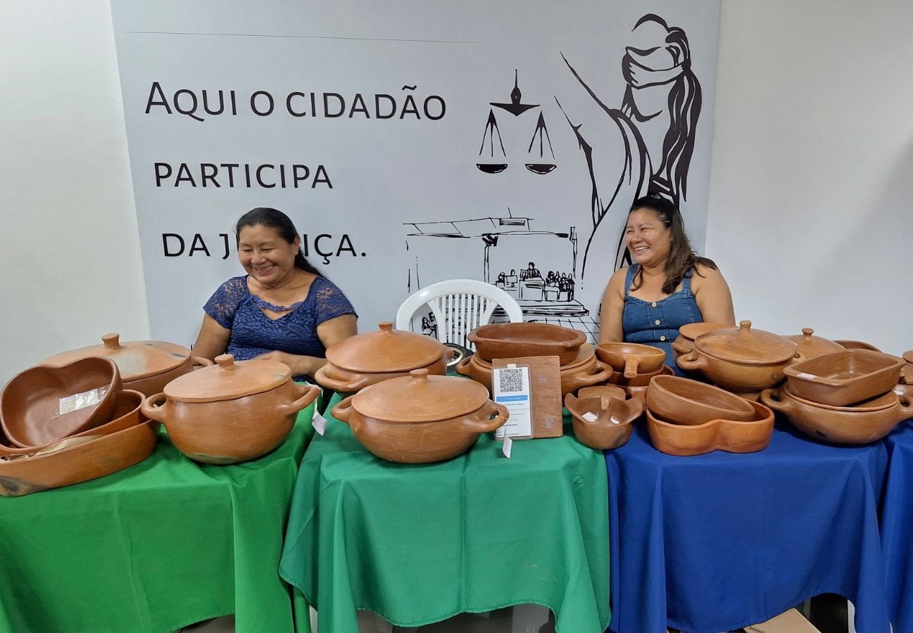 Imagem colorida mostra duas mulheres indígenas sentadas em frente a três mesas juntas ao lado uma da outra, em cima delas está diversas panelas de barro, utensílios da culinária indígena.