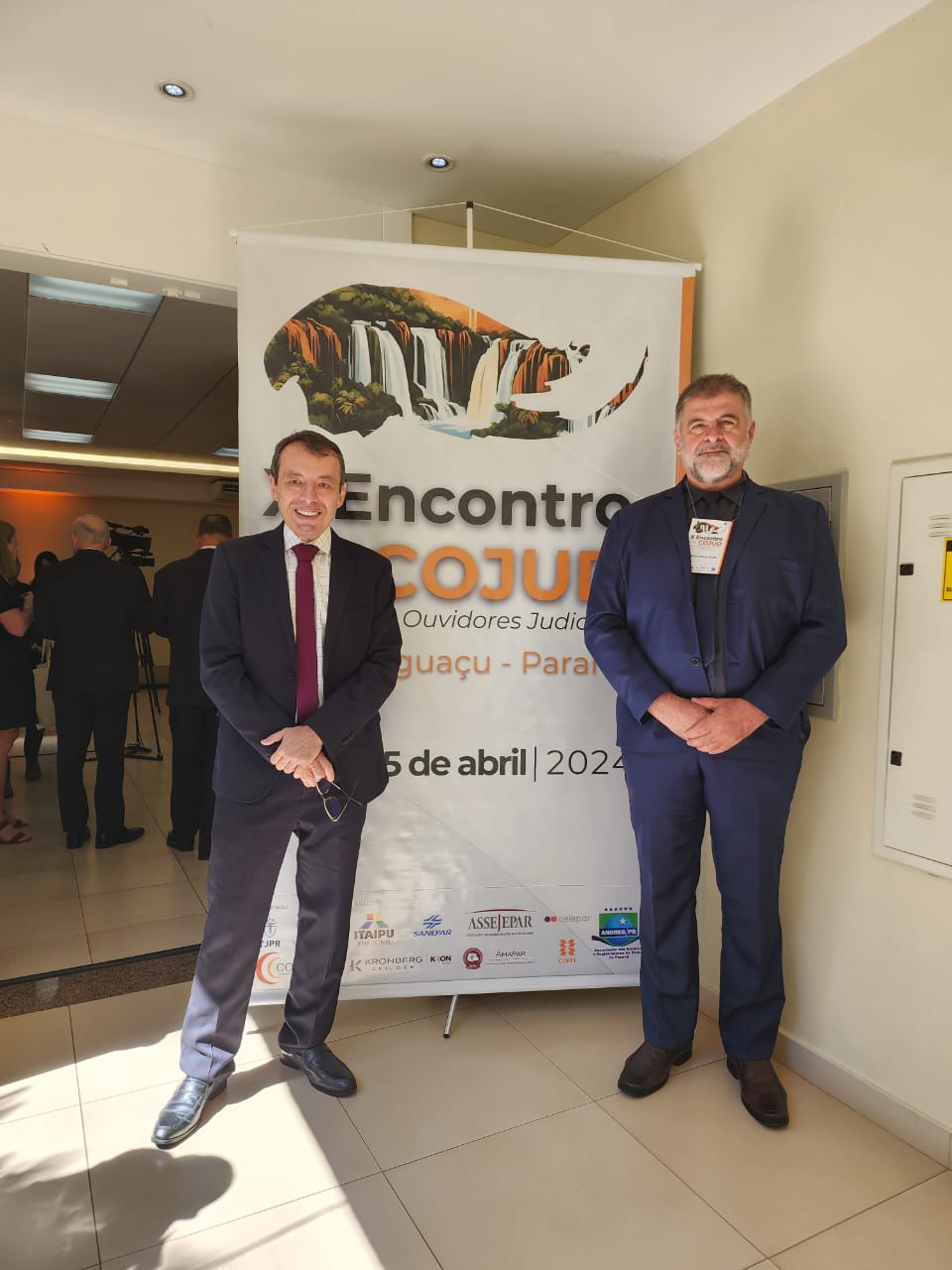 Imagem colorida mostra o desembargador Erick Linhares, e o servidor da Ouvidoria, Amadeu Triani posando para foto com o banner de divulgação do evento ao fundo.