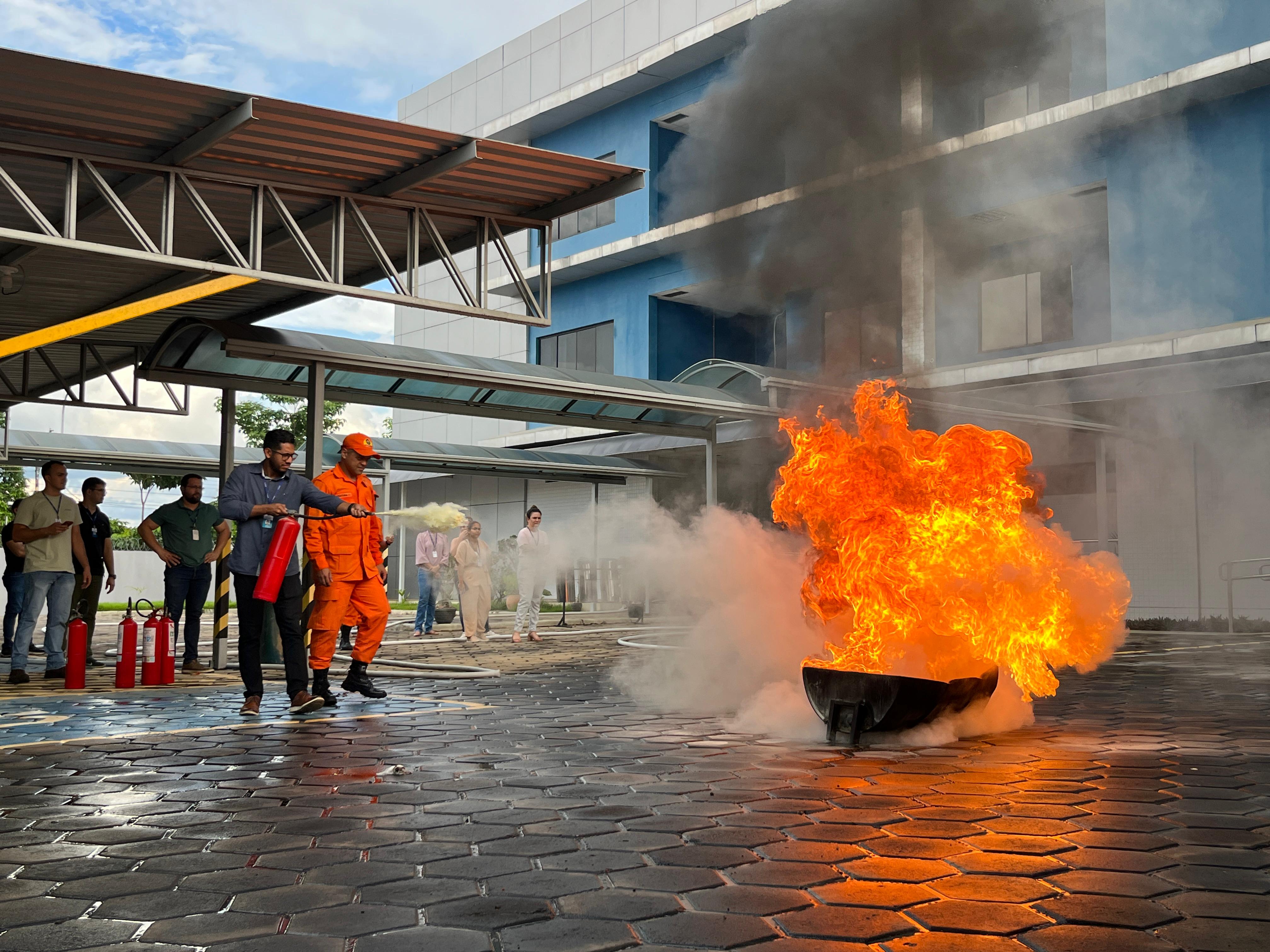 imagem colorida mostra servidores do TJRR, em área externa acompanhados de um bombeiro, durante treinamento de primeiros socorros, que ocorreu no Fórum Criminal Ministro Evancro Lins e Silva. Na imagem um dos servidores está apagando o fogo com extinto com o auxílio do Bombeiro.