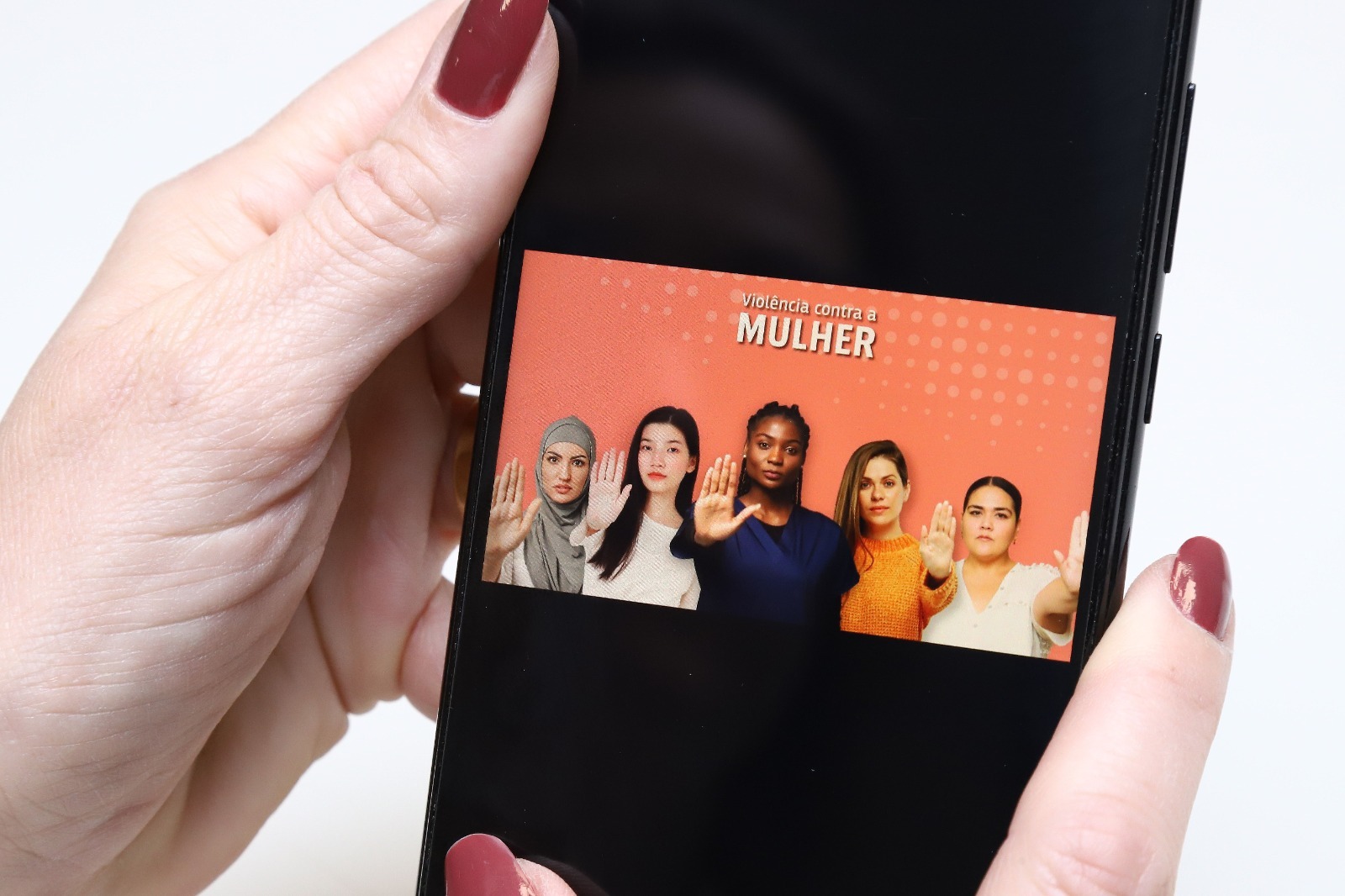 Imagem colorida mostra mão segurando um celular com o imagem com a seguinte escrita: "Proteção a Mulher", abaixo mostra cinco mulheres estendendo a mão para frente. Em alusão ao Programa EMA´NUM.